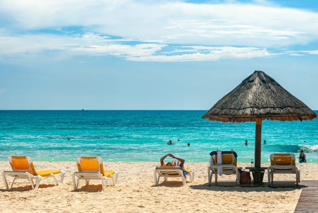 espreguiçadeiras enfileiradas com algumas pessoas deitadas e outras na praia de águas verdes cristalinas e areia amarelinha, um guarda-sol coberto com palha está ao lado, o céu é azul e há nuvens nesta praia que ilustra o post de cjip celular Cancun
