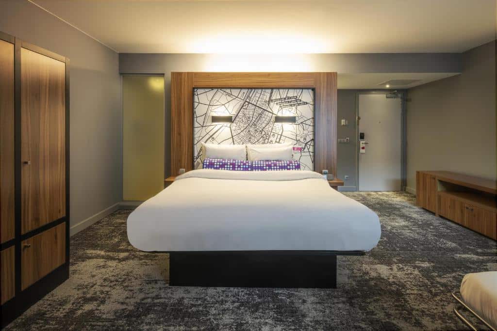 cama de casal vista de frente no Aloft Brussels Schuman com quadro na cabeceira e espaços amplos, com luzes no teto e guarda-roupa