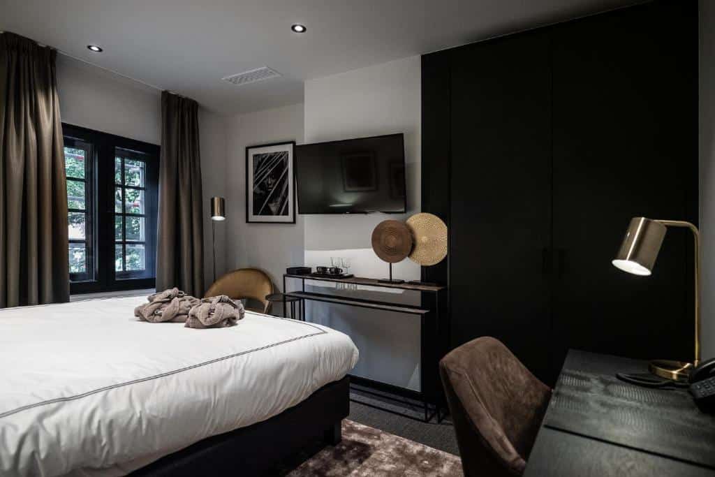 quarto do Boutiquehotel 't Fraeyhuis com cama de casal, mesa e cadeira, tv e janela de madeira, os móveis são em tons escuros e a decoração, moderna