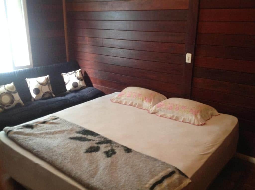 quarto com cama de casal, paredes de madeira e sofá embaixo da janela na Chacara Mairipora Essence