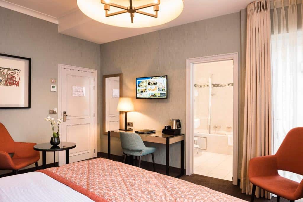 quarto do Hotel Aragon com cama de casal e detalhes laranjas com banheiro, mesa de trabalho com tv acima e, ao lado da cama, mesa e uma cadeira
