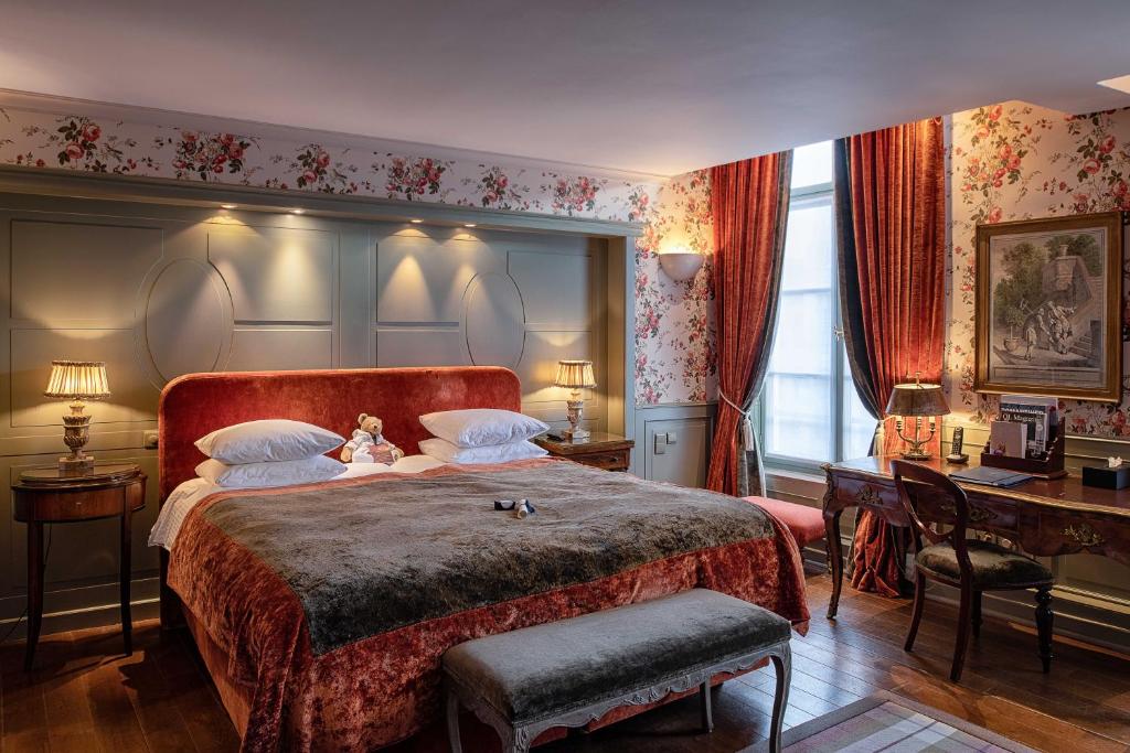 quarto amplo do Hotel De Orangerie com cama king-size e detalhes em vermelho, como cortinas e cabeceira, a janela é grande pegando toda a parede, há ainda mesa com cadeiras e luminárias ao redor da cama