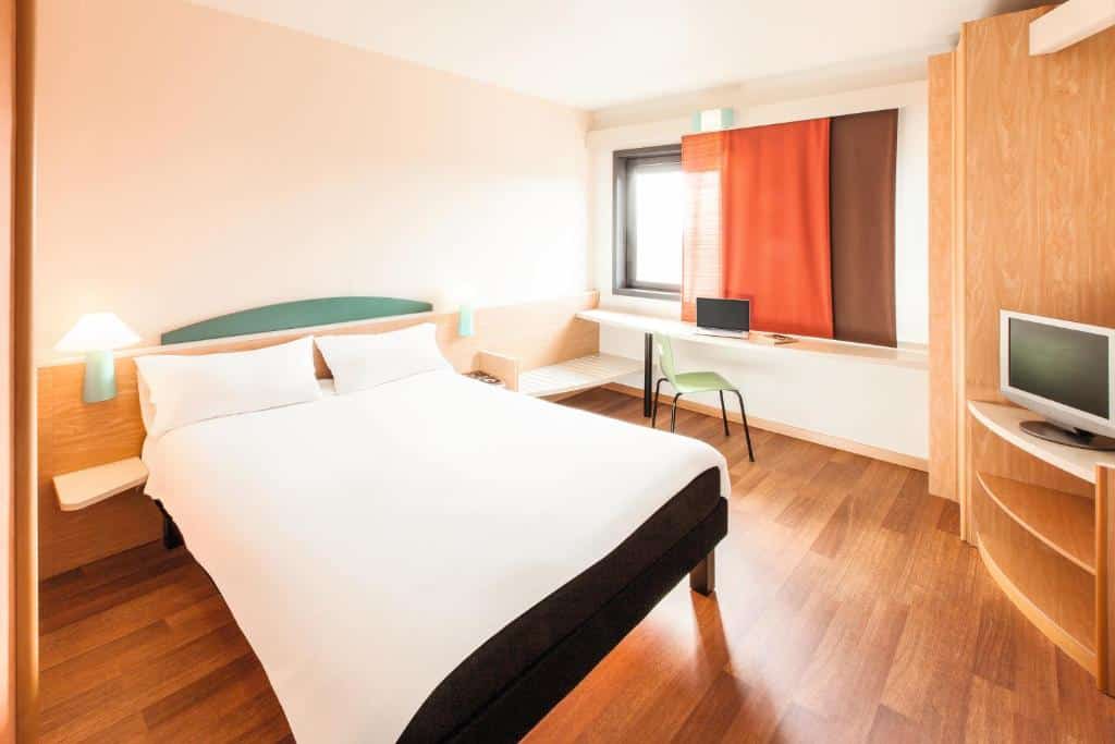 quarto espaçoso do Ibis Roma Fiera com cama de casal, janela com cortina alaranjada, guarda-roupa e tv, sendo um dos hotéis Ibis em Roma