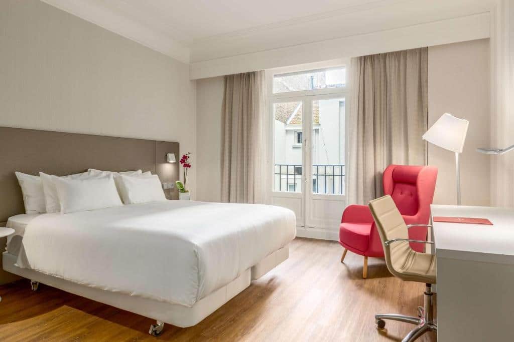 quarto do NH Collection Brussels Centre com cama de casal, poltrona vermelha e escrivaninha com cadeira em tons de branco e janela grande com cortinas