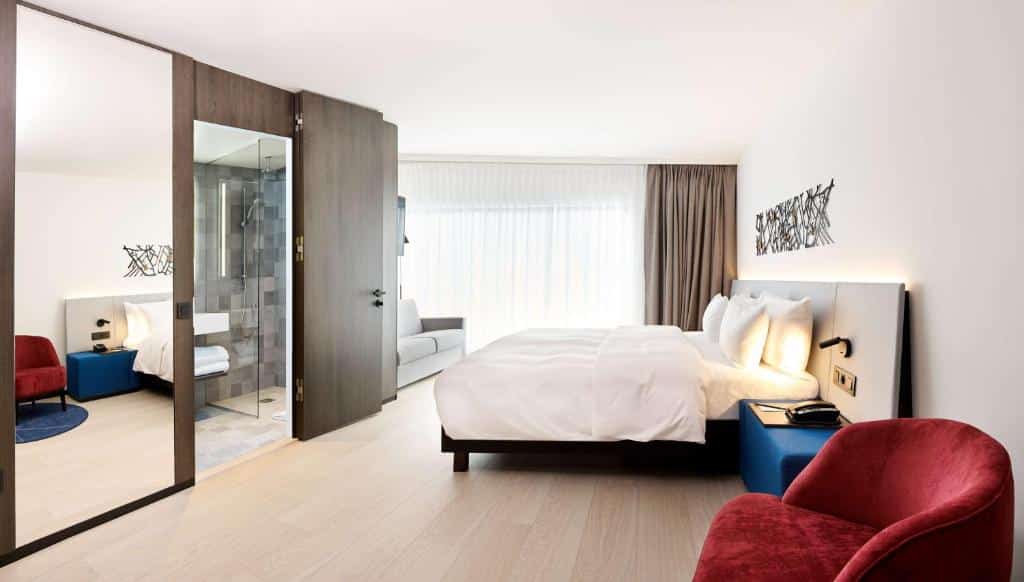 quarto amplo do Radisson Blu Hotel, Bruges com cama grande, poltrona vermelha e, do outro lado, janela enorme pegando a parede toda e um sofá, na frente da cama, há um espelho grande e entrada larga para o banheiro