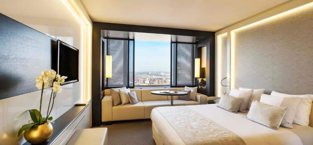 quarto amplo do The Hotel Brussels com cama de casal com tv na sua frente, janela grande que ocupa toda a parede com um sofá bem abaixo com almofadas