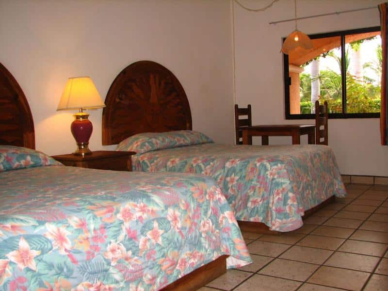 Quarto rustico do Los Barriles Hotel com duas camas de solteiro, mesa de trabalho e janela de madeira aberta.