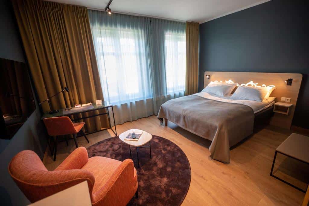 Quarto do Hotell Bondeheimen com cama de casal, poltrona laranja do lado esquerdo, no lado direito mesa de trabalho perto da janela e TV na parede em frente a cama.