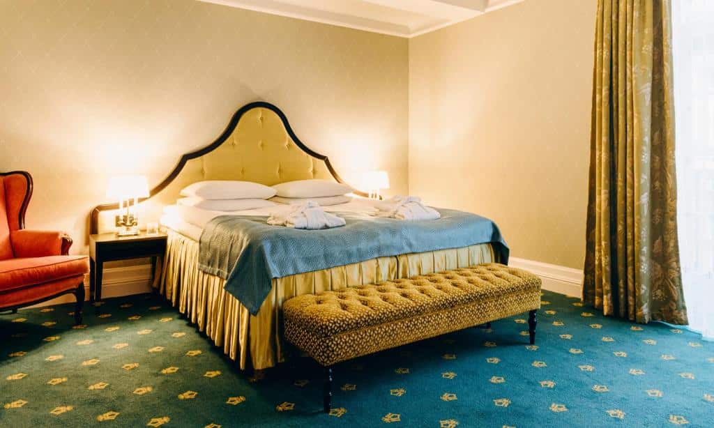 Quarto do Hotel Bristol com cama de casal com duas cômodas de cada lado da cama com luminárias e uma poltrona laranja do lado direito.