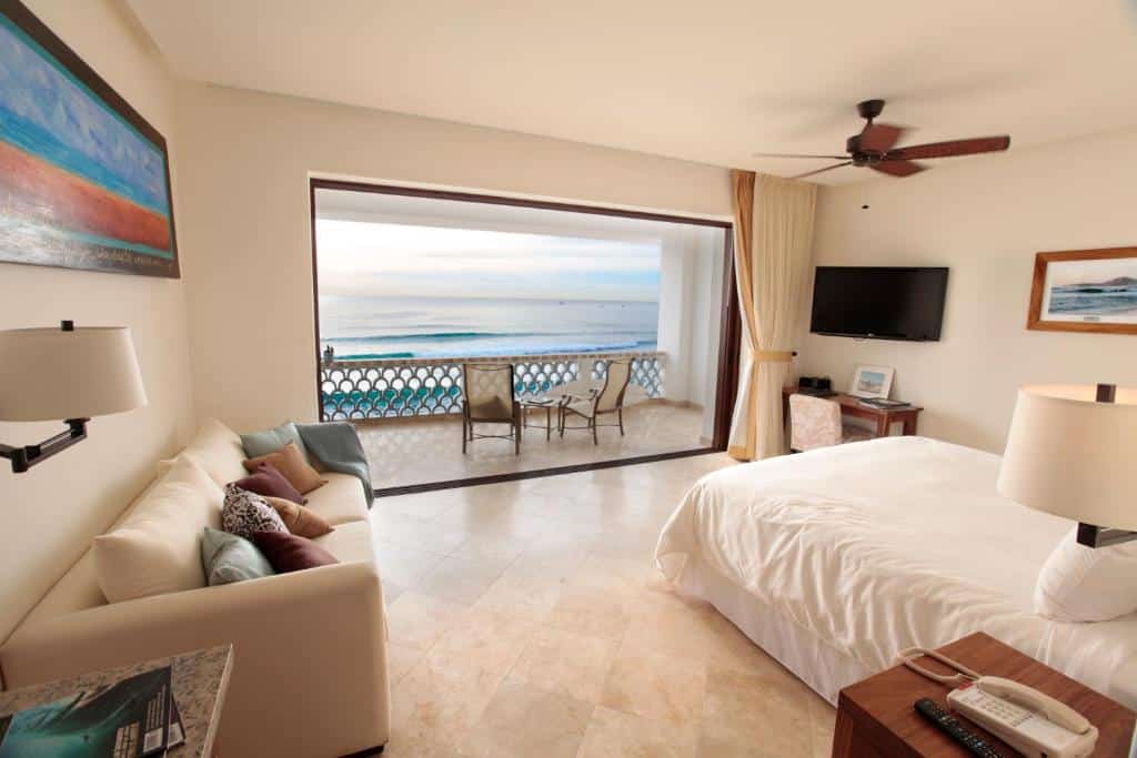 Quarto amplo do Cabo Surf Hotel com cama de casal, sofá do lado direito, TV do lado esquerdo na parede com mesa de trabalho abaixo. E em frente a cama porta ampla de vidro em frente ao mar com varanda com cadeiras.