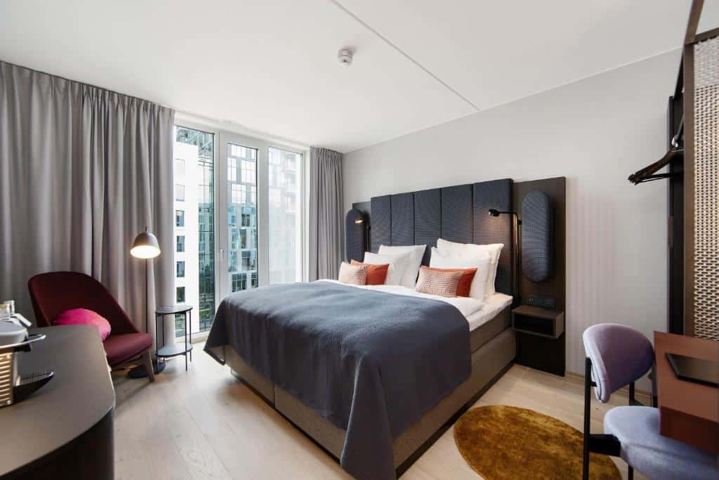 Quarto do Clarion Hotel Oslo, com cama de casal ampla, sacada, poltrona vinho do lado esquerdo e mesa de trabalho do lado direito.