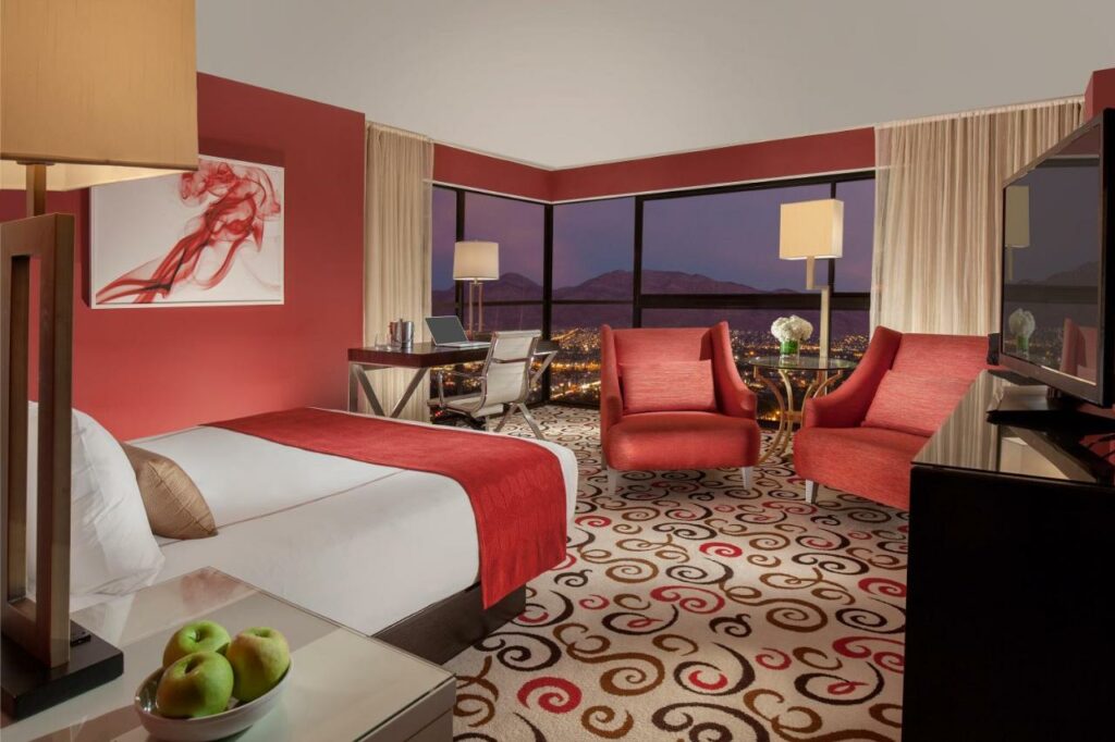 Quarto do  Downtown Grand Hotel & Casino com um carpete bege com desenhos em vermelho e marrom, uma cama de casal do lado esquerdo e uma mesinha de escritório com uma cadeira giratória, do lado direito há uma cômoda, uma televisão e duas poltronas vermelhas, atrás há uma janela panorâmico com vista para as montanhas