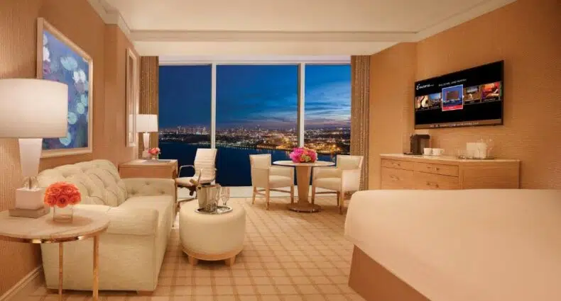 Vista do quarto Encore Boston Harbor com cama, sofá, TV e uma ampla janela com vista para a cidade de Boston. - Representa hostéis em Boston.