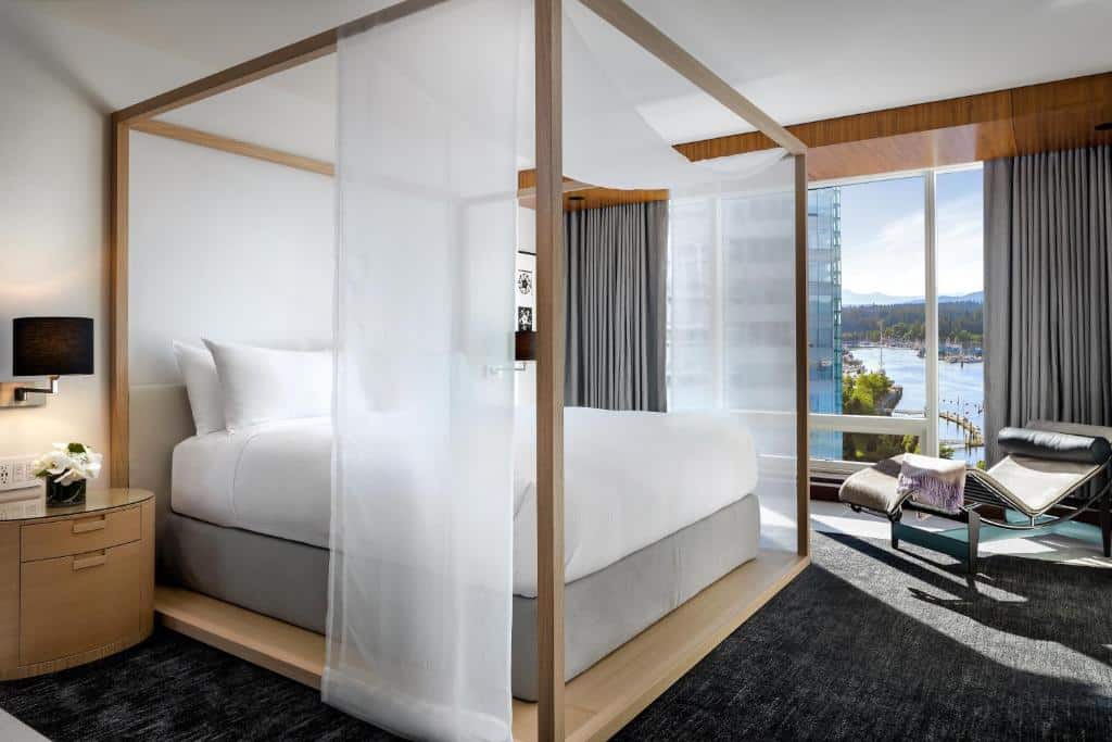 Quarto no Fairmont Pacific Rim com uma cama de casal estilo bangalô, do lado direito há uma ampla janela com vista para a baía, e do lado dessa janela há uma espreguiçadeira de couro. o chão é um carpete cinza escuro