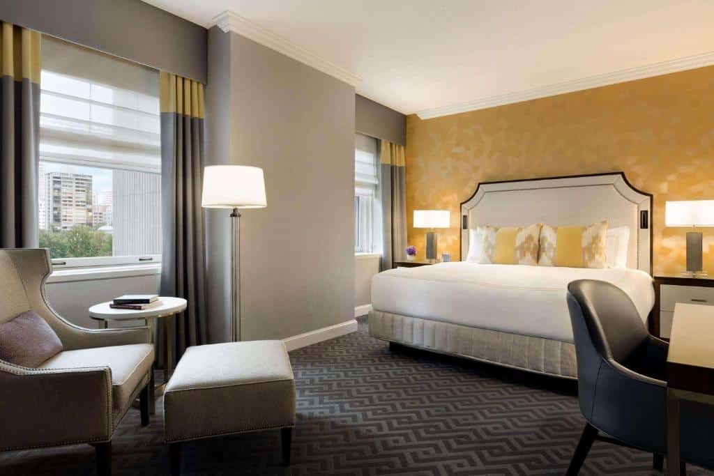 Vista do quarto do Fairmont Olympic Hotel com cama ampla, duas comodas, janelas perto, poltrona e mesa de trabalho.
