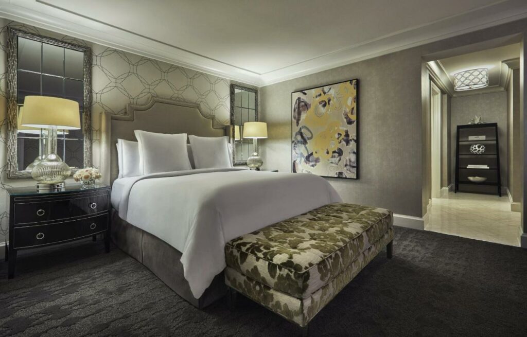 Quarto no Four Seasons Hotel Las Vegas  com uma cama de casal, uma cômoda pequena e preta do lado esquerdo da cama com um abajur, há um quadro do lado direito, o carpete é cinza escuro, o estilo da decoração é de época, para representar hotéis em Las Vegas