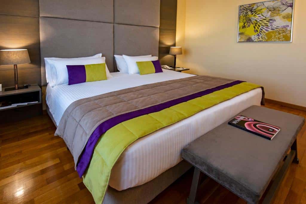 Quarto do Hotel Grand Brizo Buenos Aires com uma cama de casal ampla, chão de madeira, paredes brancas com um único quadro pendurado, duas mesinhas de cabeceira com abajur em cima, as roupas de cama são roxas, verdes e brancas