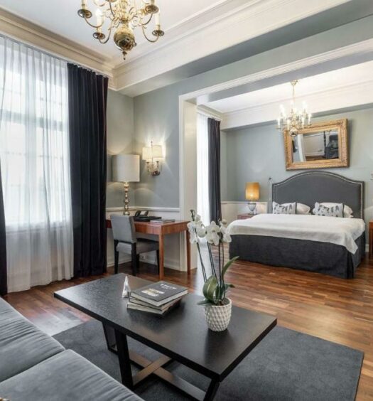 Quarto do Gran Hotel em Oslo com cama de casal, luminárias ao lado da cama, poltrona em frente da cama e poltrona na sala de estar com mesa. Representa hotéis em Oslo