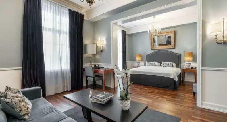 Quarto do Gran Hotel em Oslo com cama de casal, luminárias ao lado da cama, poltrona em frente da cama e poltrona na sala de estar com mesa. Representa hotéis em Oslo