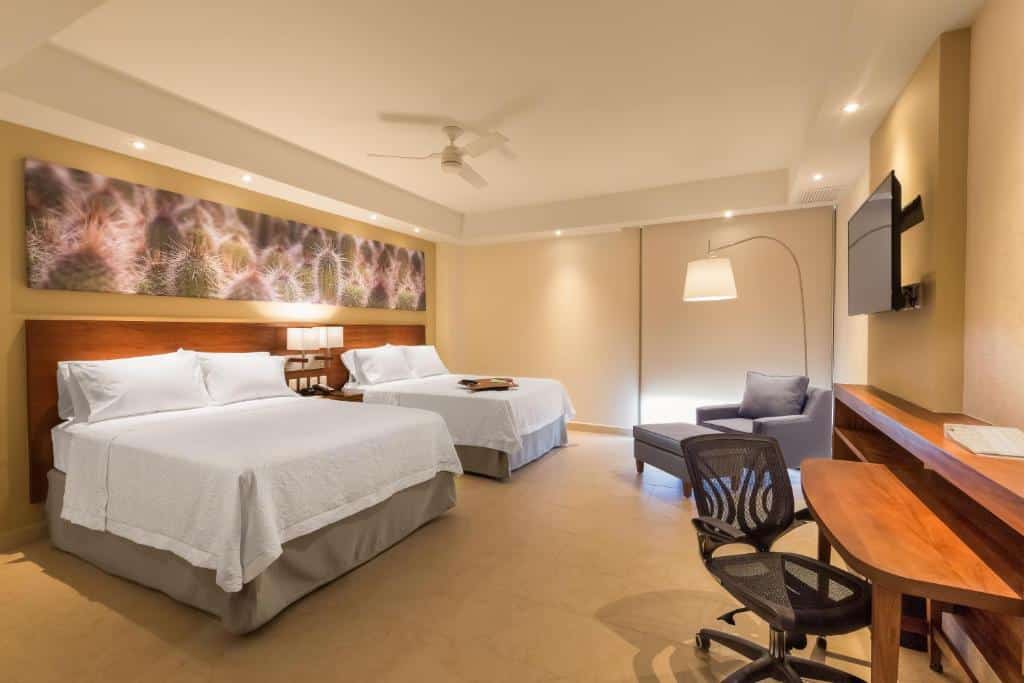 Quarto do Hampton Inn & Suites by Hilton Los Cabos com duas camas de casal e uma cômoda no meio com luminária. Poltrona cinza ao lado esquerdo. TV em frente as camas  presa na parede com mesa de trabalho e cadeira abaixo.