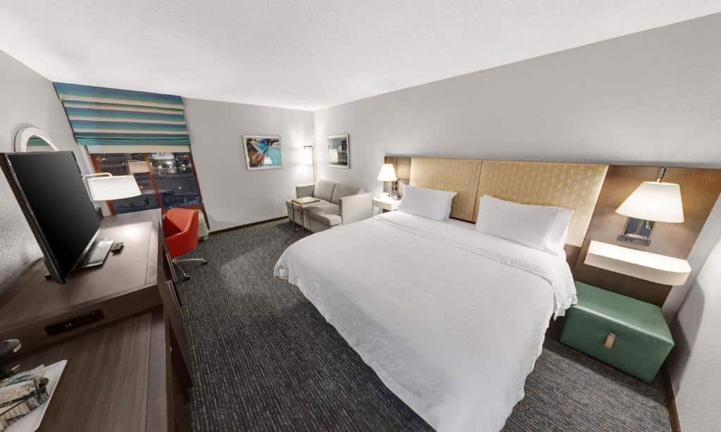 Quarto do Hampton Inn Tropicana Las Vegas com um carpete em tom de cinza e branco, uma cama de casal do lado direito, além de um pequeno sofá cinza de coisa lugares e mesinhas de cabeceira com abajures, e do lado esquerdo tem uma cômoda com uma televisão e uma janela, para representar hotéis em Las Vegas