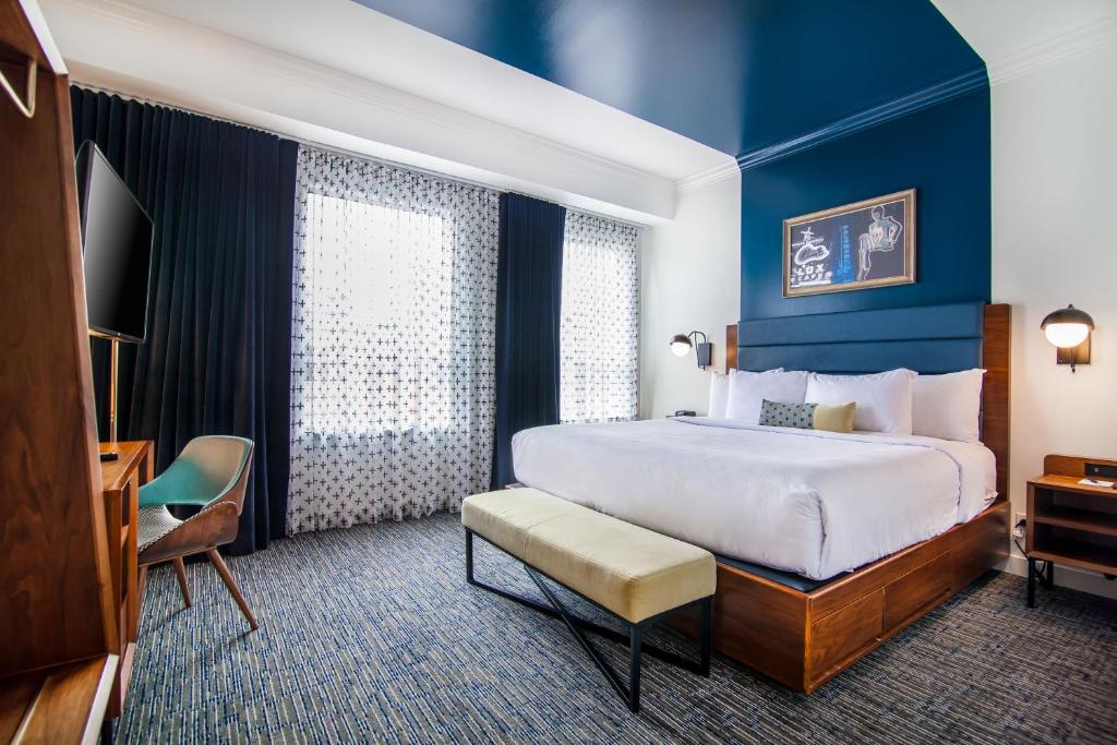 Quarto do Hotel Belmont Vancouver MGallery com uma cama de casal, duas mesinhas de cabeceira nas laterais da cama, o chão é um carpete em tons de azul e cinza, de frente para a cama há uma mesa de escritório com uma cadeira, e sob a mesa, presa na parede está uma televisão