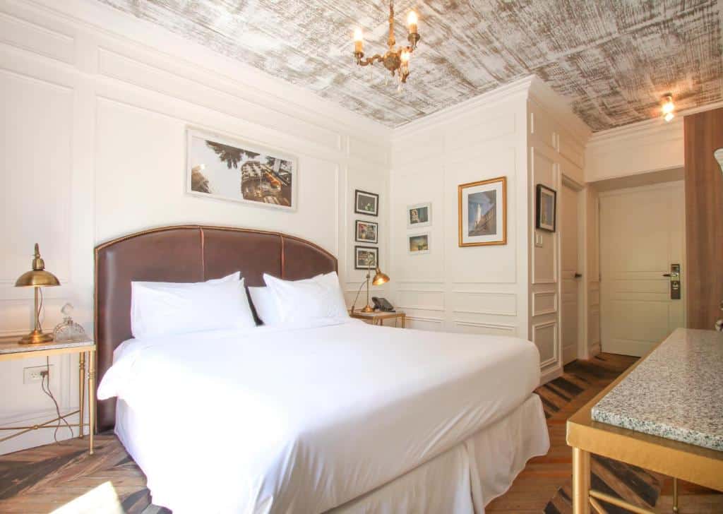 Quarto do Hotel Clasico com decoração de época, uma cama de casal, duas mesinhas de cabeceira com luminárias de ferro, alguns quadros pelas paredes brancas e o chão é de madeira