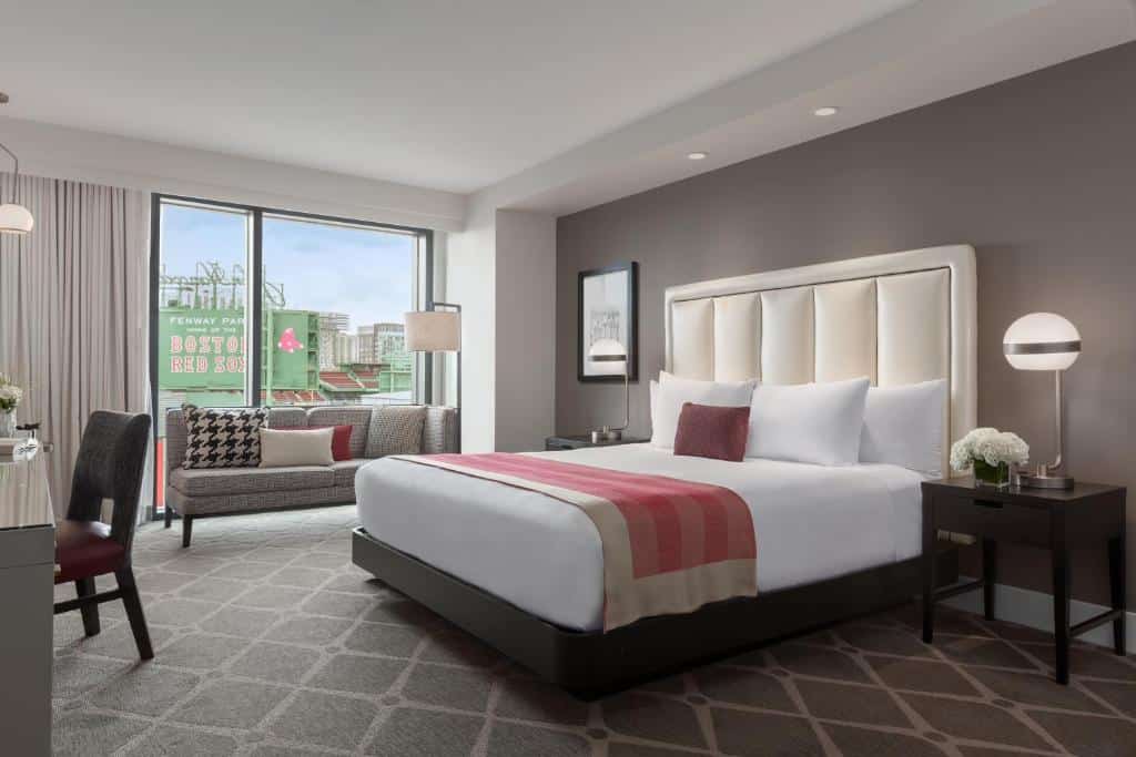 Quarto com cama ampla de casal do Boston Hotel Commonwealth com duas comodas ao redor da cama. Sofá com janela ampla e mesa de trabalho com uma cadeira.