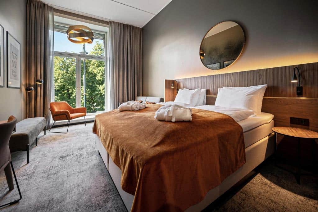 Quarto do Karl Johan Hotel com varanda, poltrona perto da sacada. A suite conta com cama de casal ampla, com duas cômodas com luminárias ao lado dela e um espelho redondo na parede em cima da cama.