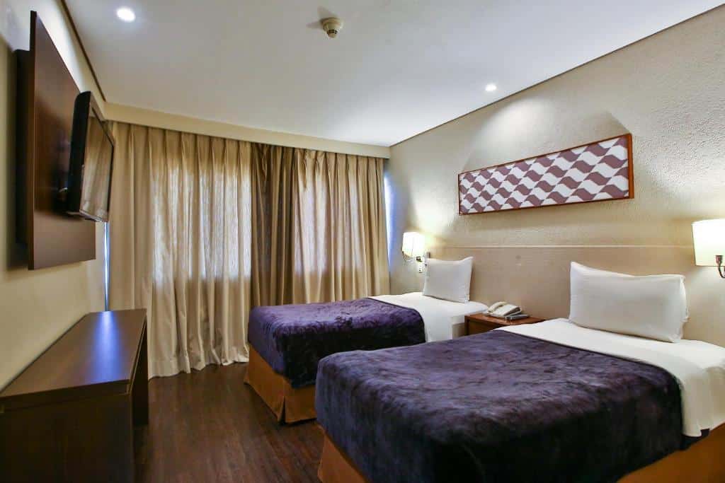 Quarto no Hotel Transamerica Berrini com uma janela com cortinas do lado das camas, há duas camas de solteiro e de frente para elas tem uma cômoda e uma televisão presa na parede
