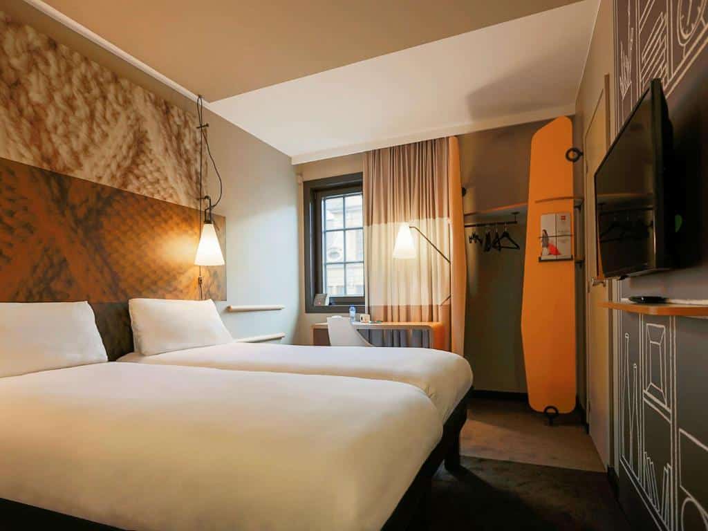 quarto com duas camas no ibis Hotel Brussels off Grand'Place, luminárias de ambos os lados da cama, detalhes em laranja e guarda-roupa com cabides e janela