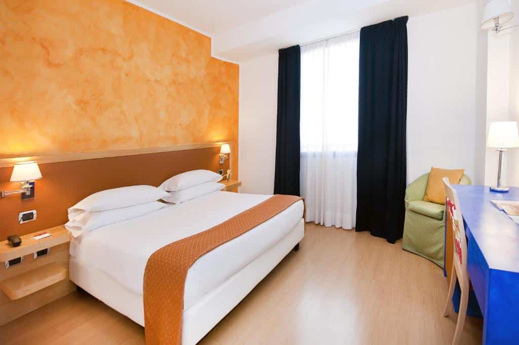 quarto amplo do ibis Styles Roma Vintage com detalhes em amarelo, na cama de casal e na parede, com janelas de cortinas escuras, poltrona e bancada