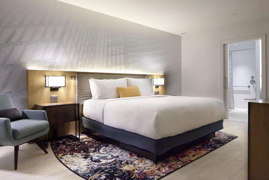 Quarto do Hotel Indigo Atlanta Downtown, com cama de casal, duas luminárias ao lado da cama com duas cômodas. Poltrona ao lado direito e porta do banheiro ao lado esquerdo.