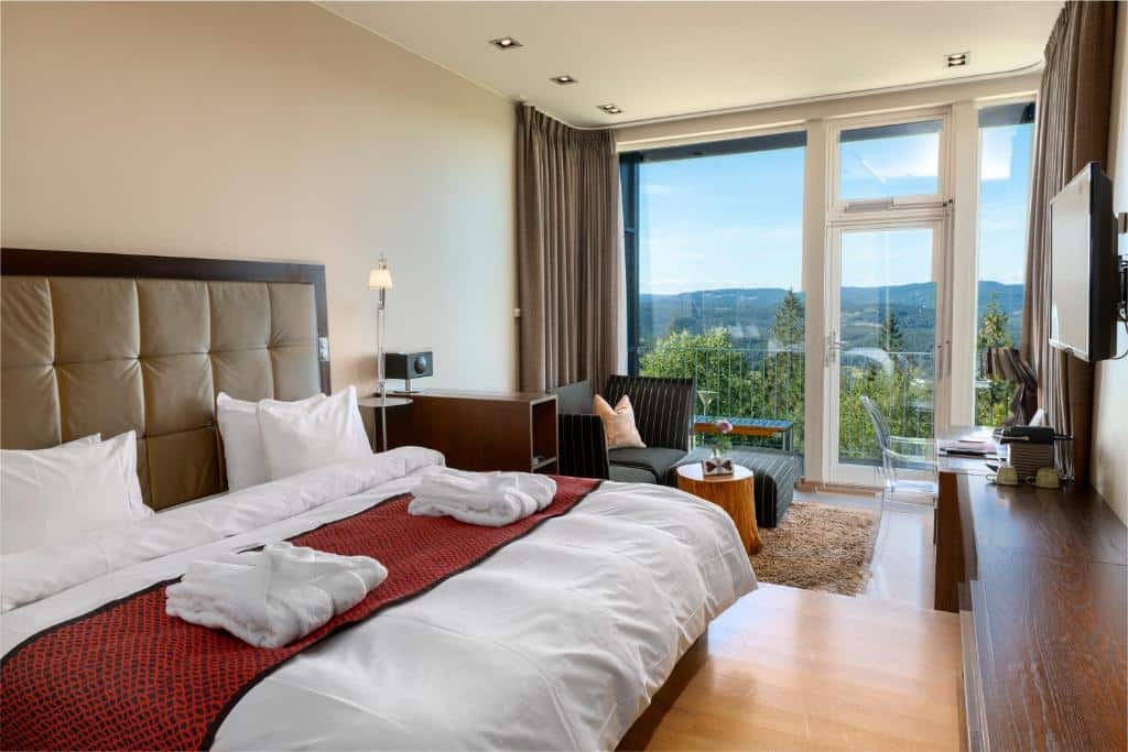 Quarto do Lysebu Hotel com cama ampla,TV a frente da cama, sofá do lado esquerdo com mesa de trabalho, e porta de vidro com sacada.