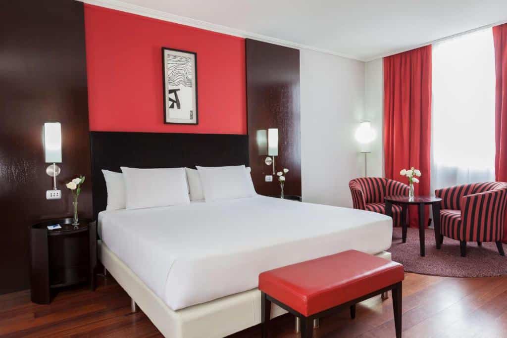 Quarto no NH City Buenos Aires com uma cama de casal, duas luminárias presas ao lado da cama, uma janela, duas poltronas e uma pequena mesinha de centro com um vaso de flor em cima, toda a decoração é em tons de preto e vermelho