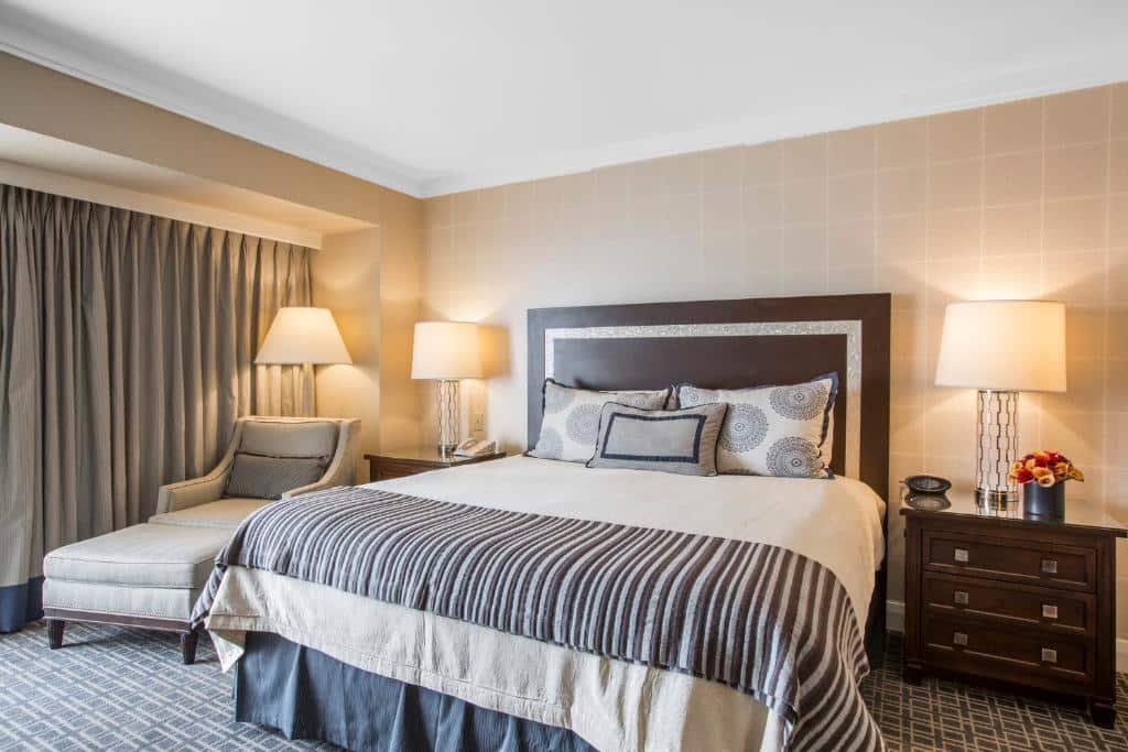 Quarto no Omni Los Angeles Hotel California Plaza com trinta e dois metros quadrados, uma cama de casal, uma pequena cômoda com um abajur em cima ao lado direito da cama, do lado esquerdo da cama há uma poltrona cinza, o chão é de carpete cinza com detalhes brancos