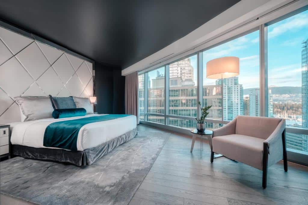 Quarto amplo no Paradox Hotel Vancouver com uma janela panorâmica com vista para a cidade, o chão é de madeira em tom de cinza, perto da janela há uma poltrona cinza e um abajur de chão ao lado, do outro lado do quarto, há uma cama de casal com roupas de cama branca e cinza