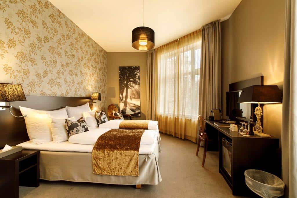 Quarto do Saga Hotel Oslo; BW Premier Collection  com tons dourados na luminária do teto, mantas da cama. O alojamento possui duas camas de solteiro, mesa de trabalho em frente as camas com TV.