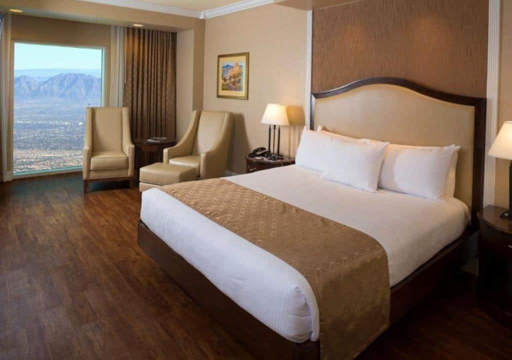 Quarto do South Point Hotel Casino-Spa com uma cama de casal, uma janela com vista panorâmica para o deserto, duas poltronas de couro bege e o chão de madeira escura
