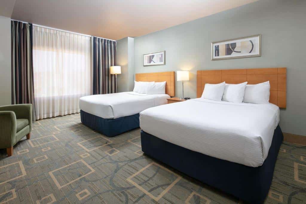 Quarto no The Platinum Hotel com uma cama de casal e uma de solteiro, o chão é de carpete cinza com desenhos geométricos em formato de quadrados, uma janela com cortinas e uma poltrona verde clara, e entre as camas há uma mesinha de cabeceira com um abajur, para representar hotéis em Las Vegas