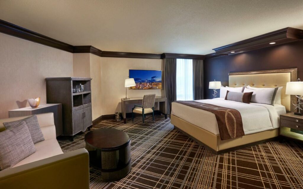 Quarto amplo no Treasure Island - TI Las Vegas Hotel & Casino, a Radisson Hotel do direito há uma cama de casal e duas cabeceiras com abajures, do lado direito da cama tem uma janela e uma mesa de escritório com uma cadeira, do lado direito do quarto há uma sofá de dois lugares e um móvel com gavetas, o chão é de carpete marrom e bege