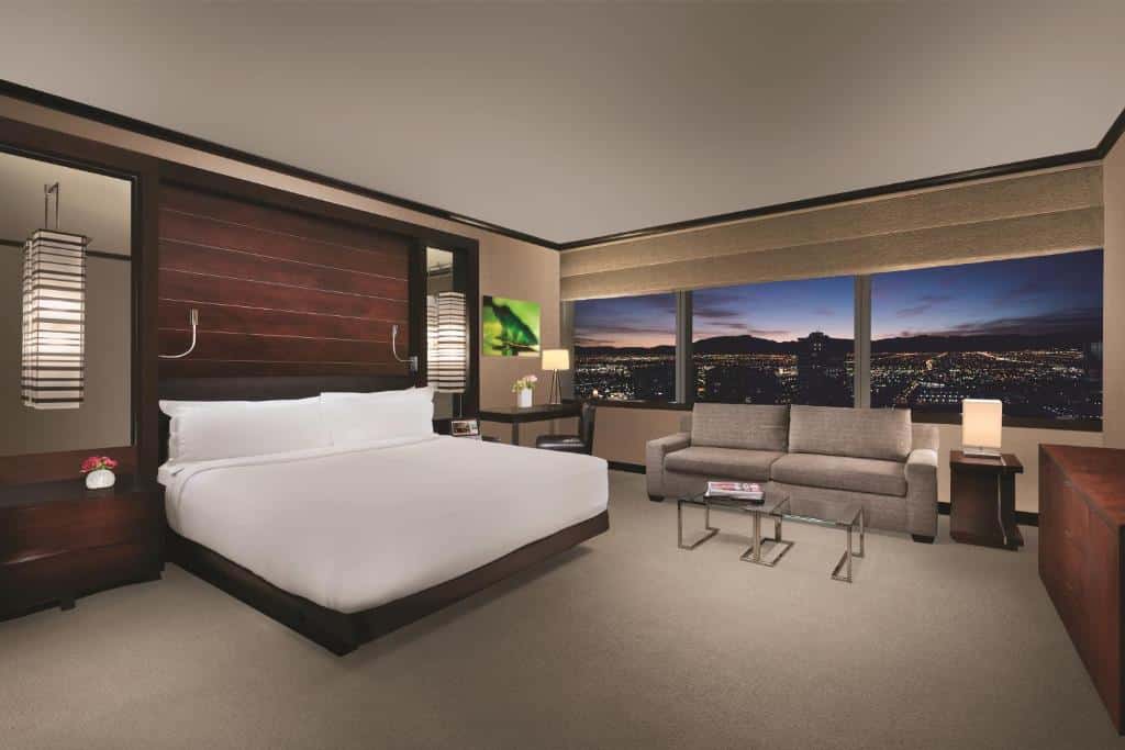 Quarto amplo no Vdara Hotel & Spa at ARIA Las Vegas com uma janela grande com vista para a cidade, com um sofá de dois lugares cinza embaixo da janela, do lado esquerdo há uma cama de casal, com duas luminárias penduradas, o chão é de carpete cinza