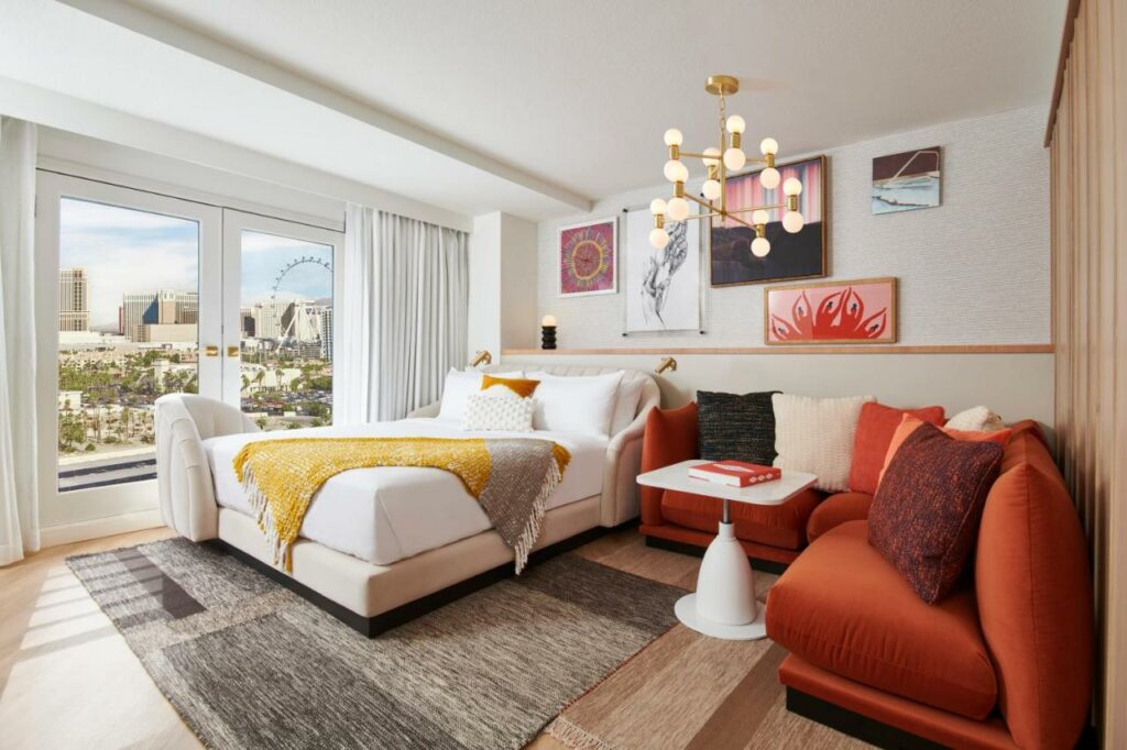Quarto do Virgin Hotels Las Vegas, Curio Collection by Hilton com uma janela ampla com cortinas brancas, uma cama de casal, um sofá em L vermelho com almofadas, um lustre com diversas lâmpadas, e alguns quadros na parede atrás da cama, o chão é de madeira clara, a decoração é em tons de vermelho, cinza e branco, para representar os resorts em Las Vegas