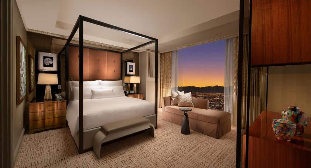 Quarto no Wynn Las Vegas com uma janela ampla do lado direito com uma sofá, do lado esquerdo tem uma cama de casal, com mesas de cabeceira com três gavetas e abajures sob elas, o carpete é em tom de bege claro, para representar resorts em Las Vegas