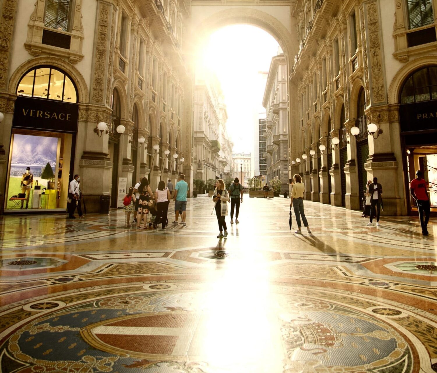 Pessoas passeando na Galleria Vittorio Emanuele II, com um raio de sol entrando no teto de vidro e refletindo no chão do local que é cheio de pinturas clássicas