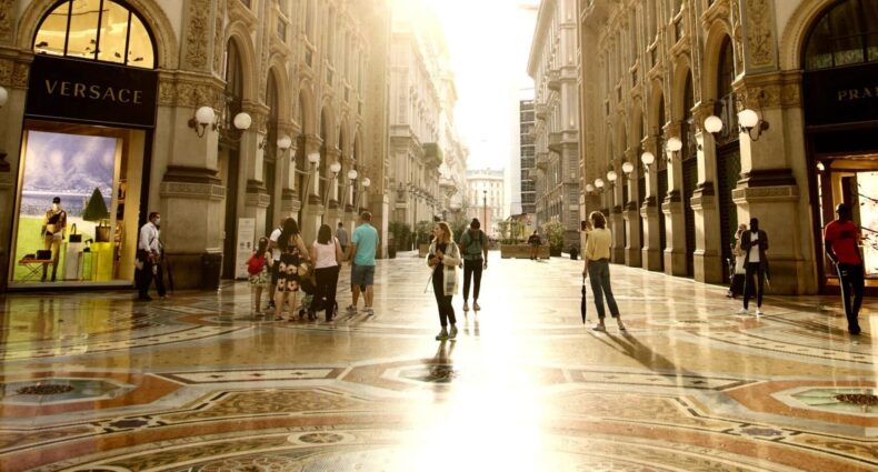Pessoas passeando na Galleria Vittorio Emanuele II, com um raio de sol entrando no teto de vidro e refletindo no chão do local que é cheio de pinturas clássicas