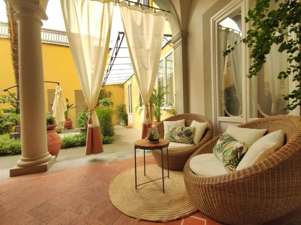 Área externa do Renascentia Garden Relais, um dos hotéis na Toscana, com 2 poltronas com 3 almofadas em cima, mesa redonda pequena ao meio, cortinas e bastante flores e jardim espalhados