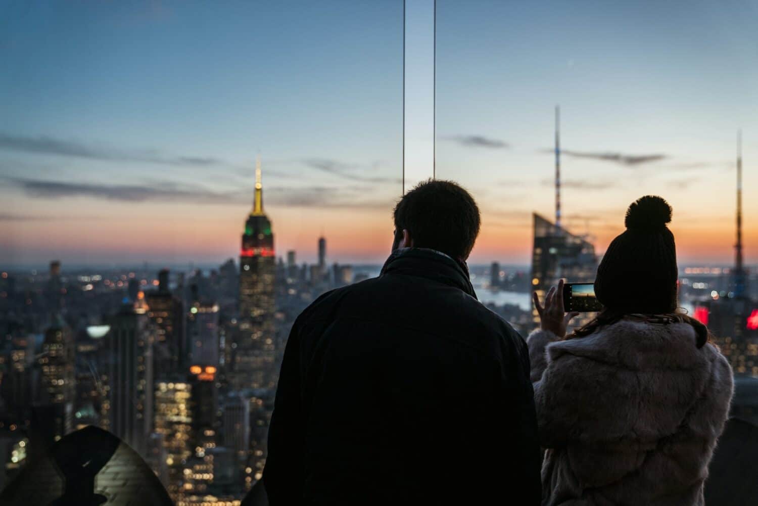 Casal olhando a cidade de Nova York, sendo a mulher segurando um celular e fotografando os prédios