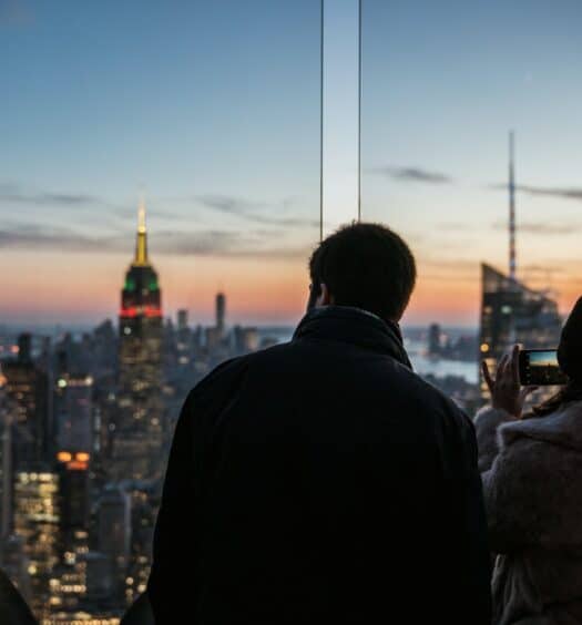 Casal olhando a cidade de Nova York, sendo a mulher segurando um celular e fotografando os prédios