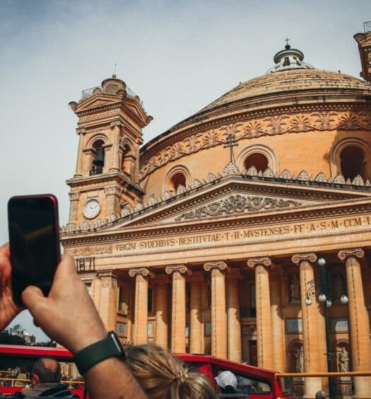 Homem usando boné e com relógio nos dois pulsos, segurando um celular preto de bordas vermelhas, para tirar foto da Rotunda de Mosta, uma igreja de Malta